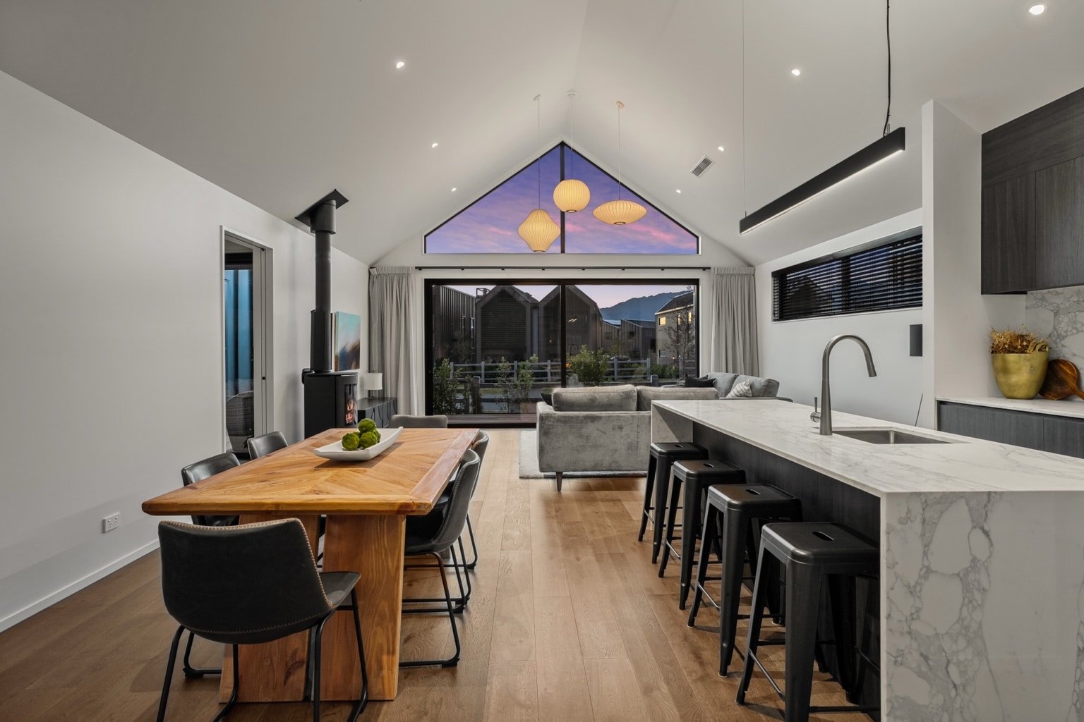 Wide plank European oak flooring in an open plan kitchen, dining and living area with marble island and gable window at twilight in a Queenstown New Zealand home