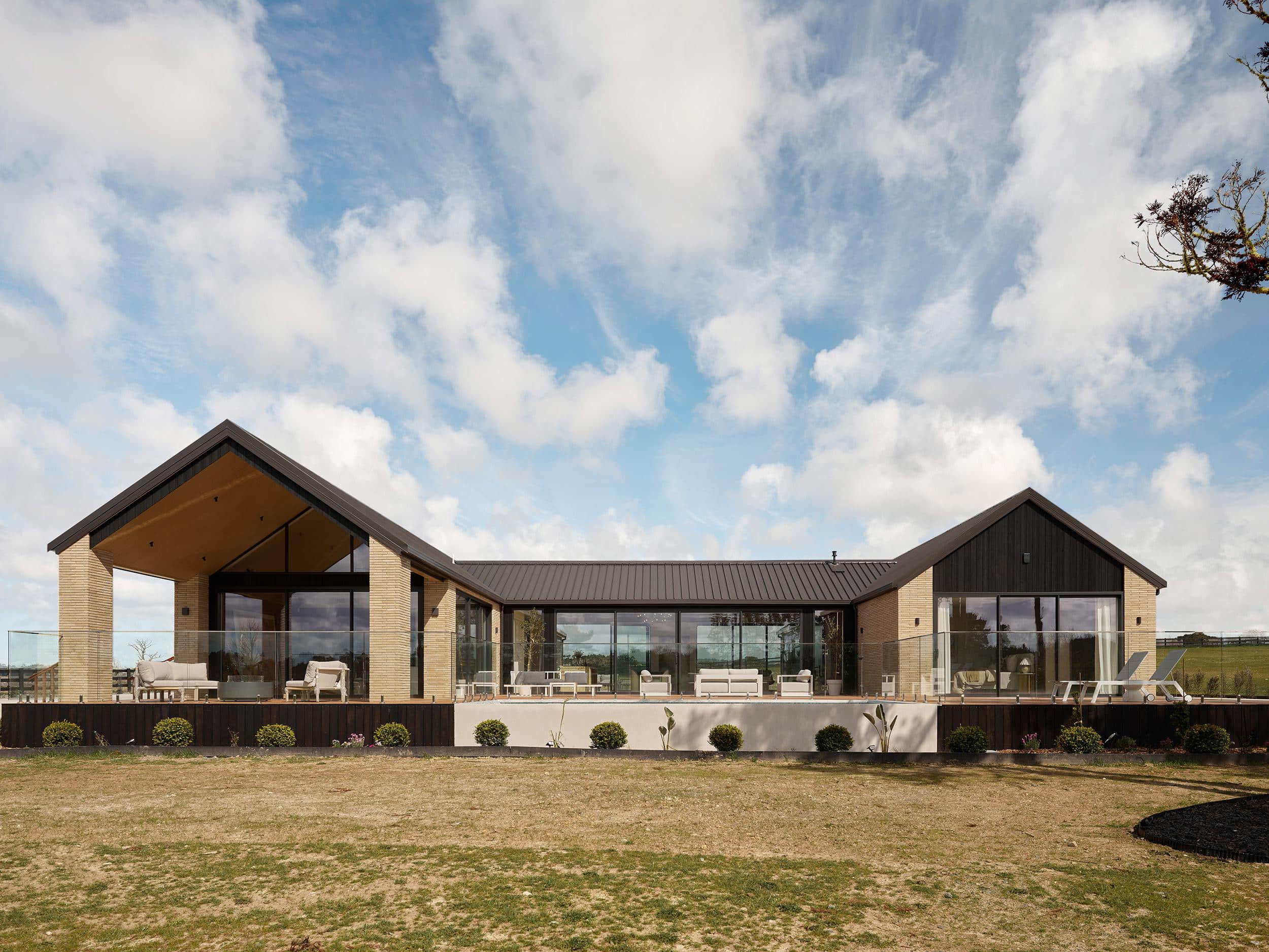 A gabled rural Auckland home in Taupaki, dark standing-seam roofs over brick and black-clad walls, set among open paddocks.