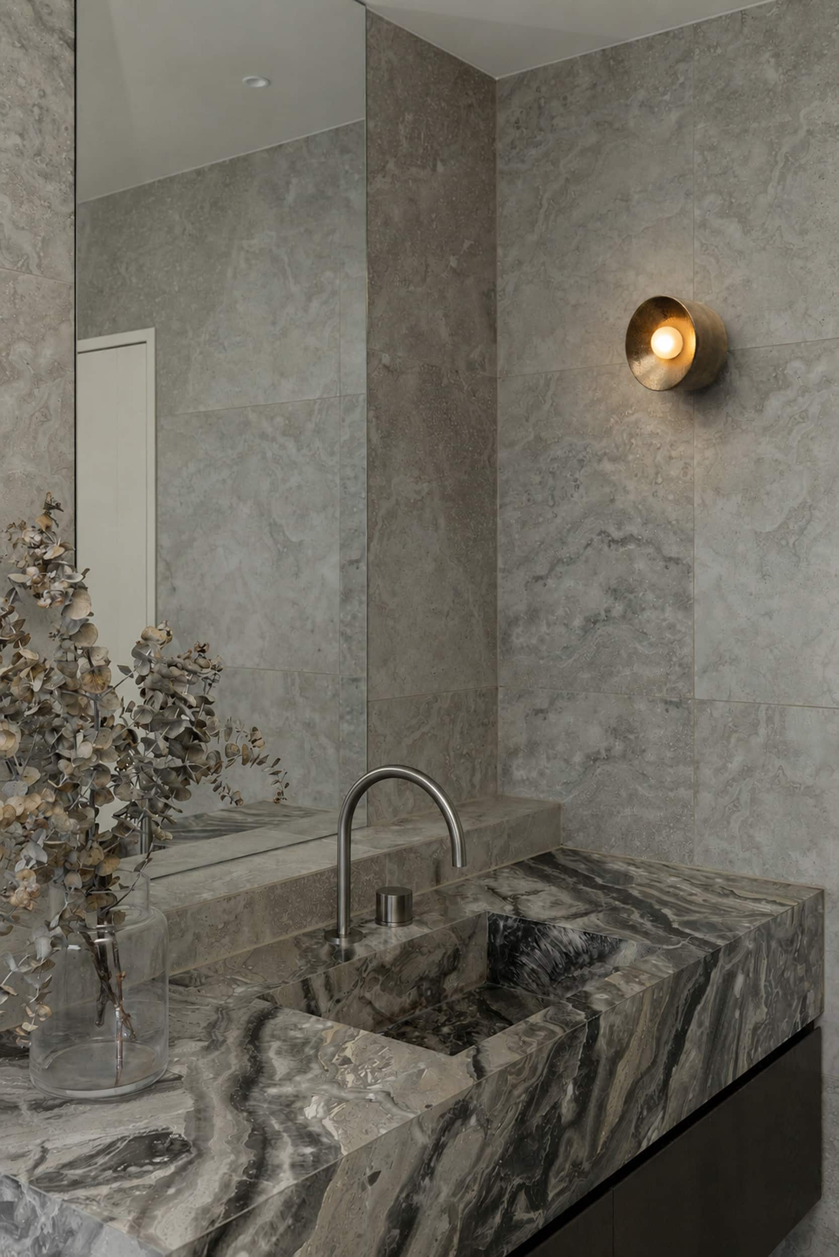 Powder room in a Hobsonville home by YG Studio, with a heavily veined stone vanity, stone tiling and a brass wall light.